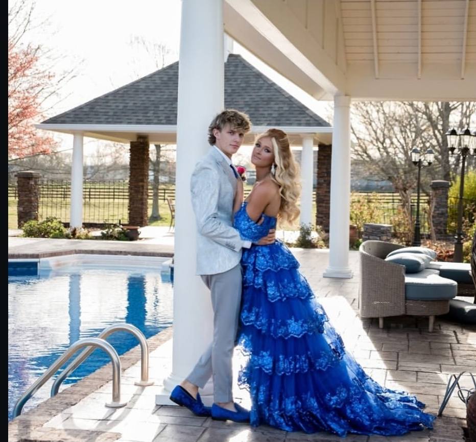 A couple posing outdoors by a swimming pool under a covered patio. The woman wears a bright royal blue tiered lace formal gown, and the man wears a light gray suit with blue shoes.