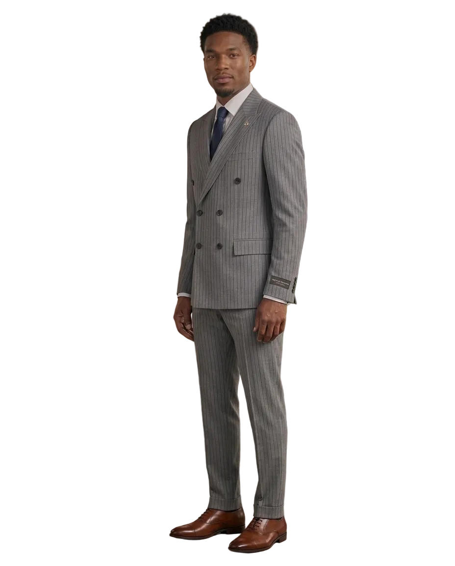 A man wearing a gray double-breasted pinstripe suit, white dress shirt, and blue tie, standing in a modern loft office setting with a leather lounge chair, wood-paneled walls, and a large window.