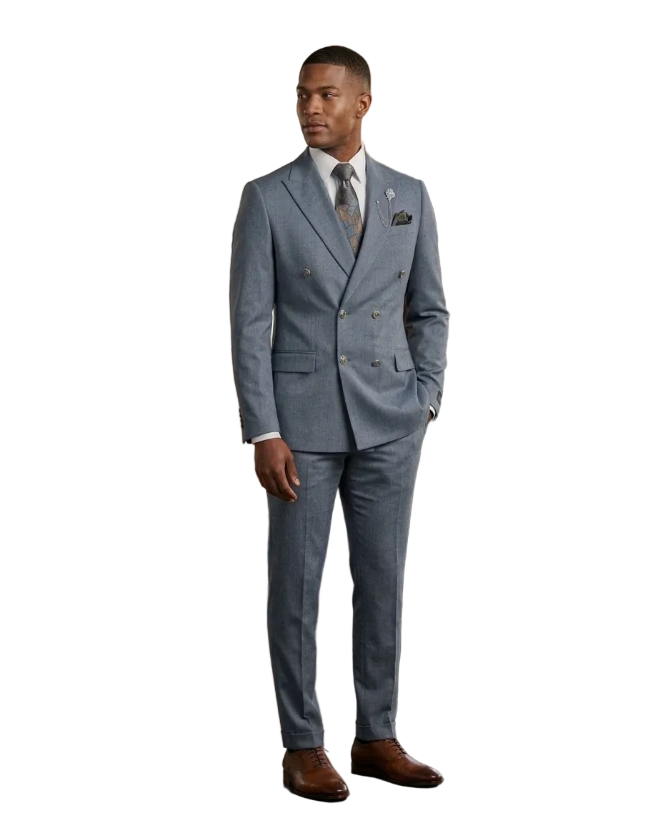 A man wearing a light gray double-breasted suit with a white shirt, patterned tie, and brown dress shoes, standing in a modern loft-style room with wood paneling, brick walls, and large windows.